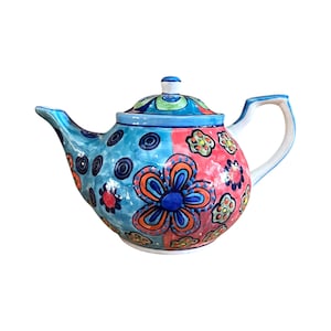 Op de afbeelding: Een handbeschilderde keramische theepot met een bloemenontwerp. De theepot heeft een blauw-roze kleurenschema, met oranje en blauwe bloempatronen. De handgreep en tuit zijn wit met blauwe accenten. Het deksel heeft een kleine knop.