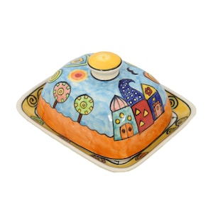 Könnte beinhalten: Handbemalte Keramik-Butterdose mit Deckel. Der Deckel zeigt eine skurrile Landschaft mit Häusern, Bäumen und blauem Himmel. Die quadratische Basis hat einen gelben Rand und dekorative Wirbel.