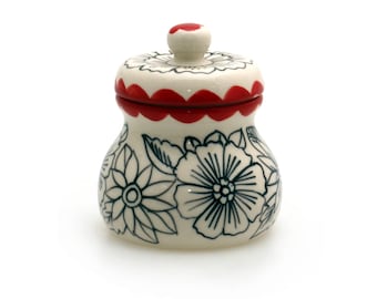 May include: A white ceramic jar with a red and white lid. The jar is decorated with black floral designs.