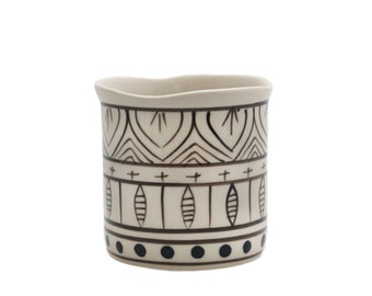 May include: A white ceramic cup with a black and white geometric pattern. The pattern includes a series of lines, dots, and shapes.