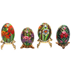 Può includere: Quattro uova di Pasqua decorative dipinte a mano su supporti in ottone. Ogni uovo presenta un design floreale unico in colori vivaci, tra cui rosso, verde, blu e rosa. Le uova sono esposte su uno sfondo bianco, evidenziando i dettagli intricati.