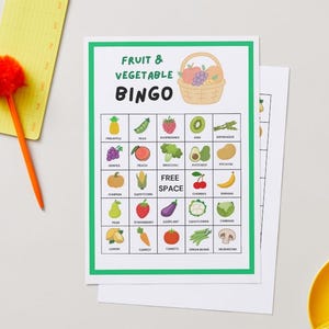 May include: A Fruit & Vegetable Bingo game card with a green border, featuring illustrations of various fruits and vegetables. The card includes a basket of fruit graphic and the words "Fruit & Vegetable Bingo". A yellow notepad and orange pen are on the left.