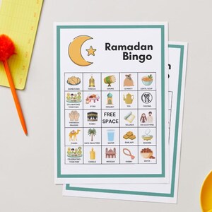 May include: Two Ramadan Bingo game cards with a teal border, featuring illustrations of Ramadan-related items. The words "Ramadan Bingo" are at the top. A yellow notepad and orange pen with a pom-pom are on the left. A yellow teacup is on the right.