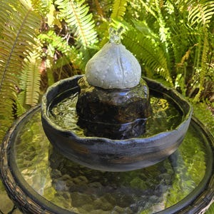 May include: A small outdoor water fountain with a gray ceramic bowl and a decorative top. The fountain sits on a glass-topped table with a woven base, surrounded by green foliage.