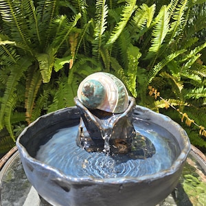 May include: A dark gray ceramic water fountain featuring a large, iridescent green and white seashell at the center. Water flows from the shell into a wide, circular basin with a textured, uneven rim, creating ripples on the surface of the water.