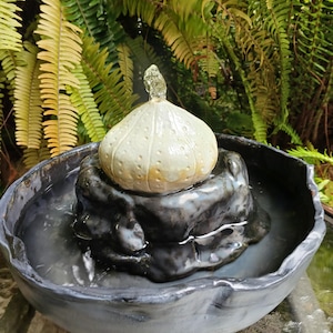 May include: A small, ceramic water fountain with a light beige, bulb-shaped top and a dark gray base. Water flows from the top into a dark gray bowl. The fountain is set against a backdrop of green ferns.