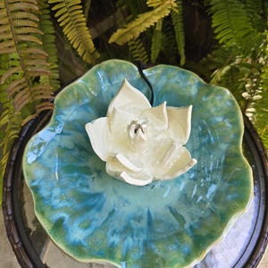 May include: A ceramic water fountain featuring a white lotus flower centerpiece resting in a wide, scalloped basin with a glossy, blended glaze of turquoise, teal, and green. A thin black power cord extends from the center of the white flower.