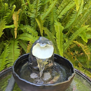 May include: A dark gray ceramic water fountain featuring a central sculpture with a white, shell-like opening. Water flows from the center piece into a wide, circular basin. The fountain has a glossy finish and is filled with clear, moving water.