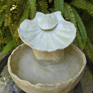 May include: A small, two-tiered outdoor fountain with a seashell design. The fountain is made of a light green ceramic material and features water cascading from the top shell into the lower basin. The fountain is set on a glass table with a natural green background.