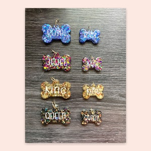 May include: Eight dog bone-shaped pet ID tags with glitter embedded in the resin. The tags are in various colours, including blue, purple, gold, and black. The names "Royal", "Jewel", "King", and "Queen" are written on the tags.