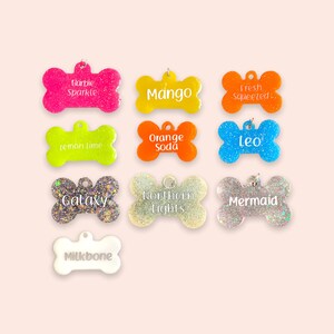 May include: Ten colourful dog bone-shaped tags with glitter and white text. The tags are in various colours, including pink, yellow, orange, blue, and silver. The text on the tags includes "Barbie Sparkle", "Mango", "Fresh Squeeted", "Lemon Lime", "Orange Soda", "Leo", "Galaxy", "Northern Lights", "Mermaid", and "Milkbone".