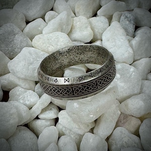 Może przedstawiać: Srebrny pierścień z teksturowaną powierzchnią i skomplikowanymi wzorami celtyckich węzłów i run. Pierścień jest umieszczony na tle białych kamieni, podkreślając jego szczegółowe wykonanie. Wyrazisty dodatek.