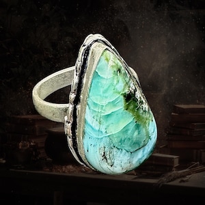 May include: A silver ring featuring a large, teardrop-shaped stone with a smooth, polished surface. The stone displays a gradient of vibrant turquoise and seafoam green colors with dark, organic veining patterns throughout the center.