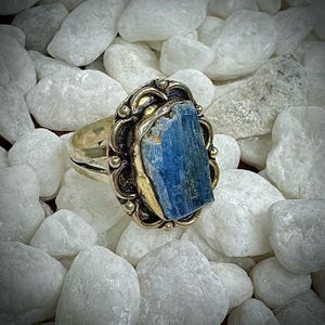 Puede incluir: Un anillo de plata adornado con una gran piedra de cianita azul rectangular. La piedra está engastada en un bisel dorado con detalles decorativos. El anillo se exhibe sobre un lecho de piedras blancas.