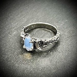 May include: A silver ring featuring a light blue oval gemstone. The ring has a detailed, textured band with a crescent moon design. The gemstone is set in a decorative setting, adding to the ring's unique aesthetic.