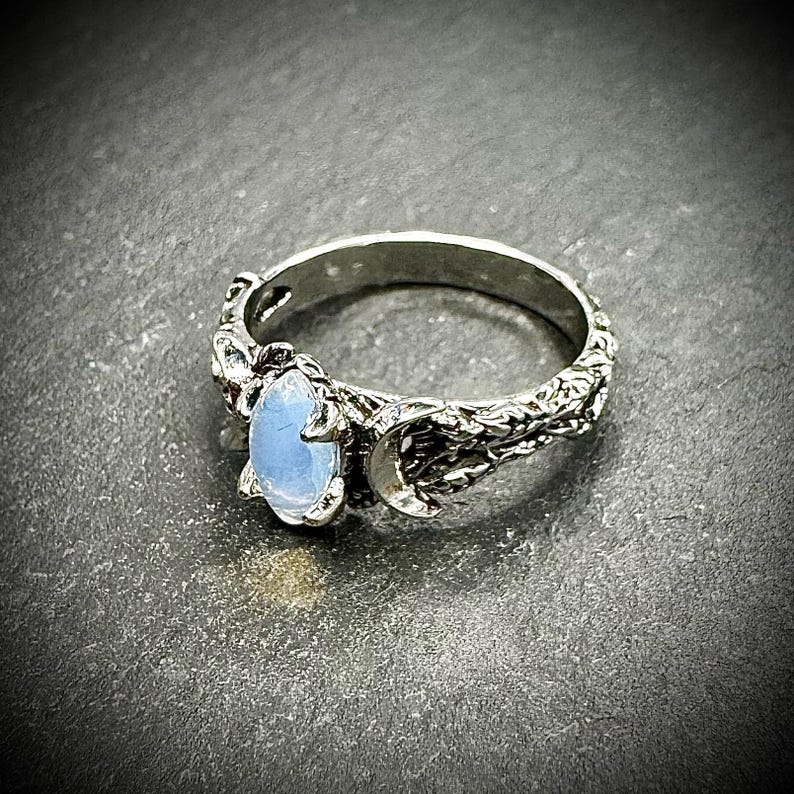May include: A silver ring featuring a light blue oval gemstone. The ring has a detailed, textured band with a crescent moon design. The gemstone is set in a decorative setting, adding to the ring's unique aesthetic.
