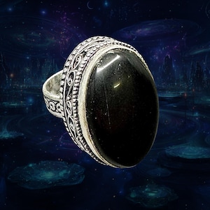 May include: A silver ring with an oval, black gemstone. The ring has an ornate, detailed design around the gemstone and on the band. The gemstone is smooth and polished, reflecting light. The ring is set against a dark, space-themed background.