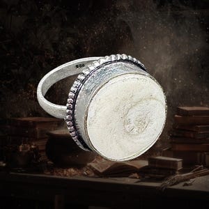 Peut inclure: Une bague en argent avec une grande incrustation circulaire d'ammonite fossilisée blanc cassé. L'anneau en métal présente une finition texturée et oxydée avec une bordure crantée décorative entourant la pierre centrale. L'intérieur est marqué 925.