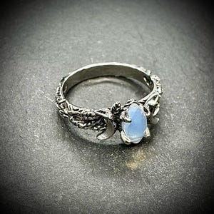 May include: A silver ring with an oval, light blue gemstone. The ring features a detailed, nature-inspired design with leaves and a crescent moon. The band has an intricate, textured pattern.