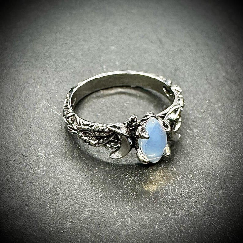 May include: A silver ring with an oval, light blue gemstone. The ring features a detailed, nature-inspired design with leaves and a crescent moon. The band has an intricate, textured pattern.