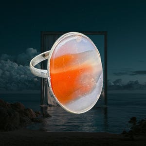May include: A silver-toned ring featuring a large, oval-shaped cabochon stone. The stone displays a smooth, polished surface with swirling patterns of translucent white and opaque, vibrant orange colors set within a simple metal band.