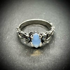 May include: A silver ring featuring a light blue, oval-shaped gemstone. The ring has a detailed design with crescent moon and leaf accents on the band. The ring is set against a dark gray background.