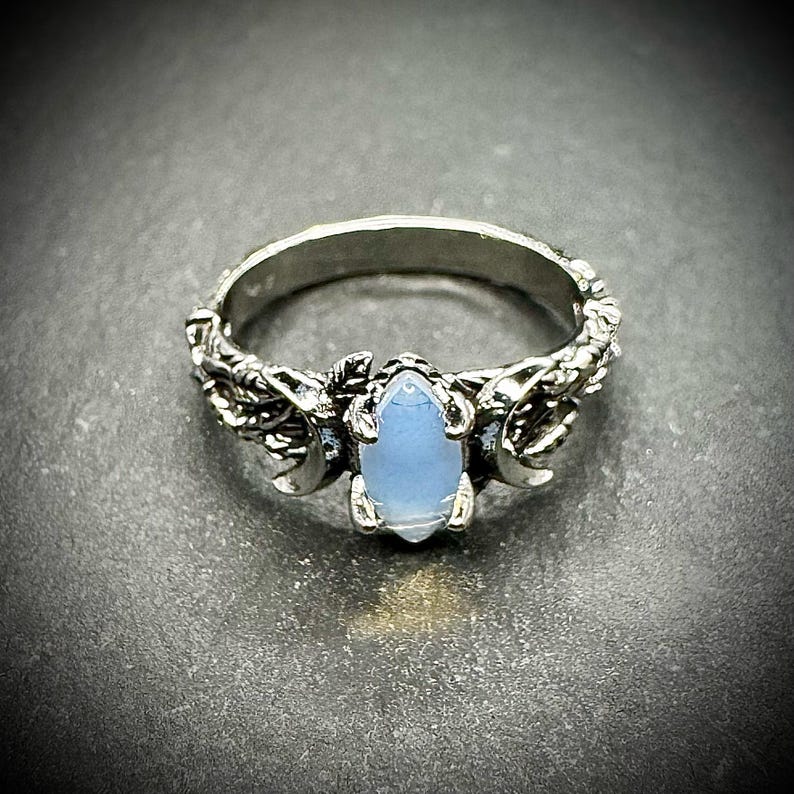 May include: A silver ring featuring a light blue, oval-shaped gemstone. The ring has a detailed design with crescent moon and leaf accents on the band. The ring is set against a dark gray background.