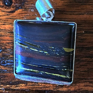 May include: A square pendant with a silver-colored bail. The pendant features a polished stone with a mix of dark blue, brown, and gold colors. The stone has a layered, marbled appearance, and is set in a silver-colored metal frame.