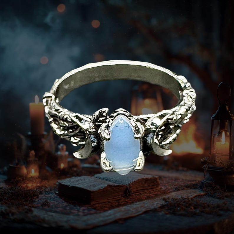May include: A silver-toned ring with a light blue, teardrop-shaped gemstone. The ring features ornate, textured detailing around the band and the setting of the stone. The background includes candles and an open book, creating a mystical aesthetic.