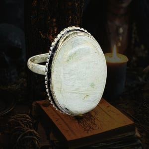 May include: A silver-toned ring featuring a large, oval-shaped, milky white stone set in a decorative, textured metal bezel. The band is simple and metallic, supporting the prominent, smooth, light-colored cabochon stone that dominates the design.