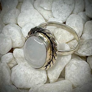 May include: A silver ring with a large, oval, pale blue gemstone. The gemstone is set in a decorative bezel with leaf-like details. The ring band is smooth and polished. The ring is displayed on a bed of white stones.