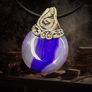 Puede incluir: Un collar con colgante que presenta una piedra grande, pulida y de forma ovalada con bandas de color púrpura, blanco y gris. La piedra está rematada con una tapa de metal dorado con un diseño ornamentado. El collar cuelga de un cordón negro.
