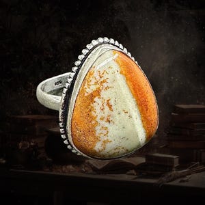 May include: A silver ring with a large, teardrop-shaped stone. The stone is a mix of orange, white, and tan. The ring has a textured bezel setting and a smooth band. The ring is set against a dark background. The ring is marked with '925'.
