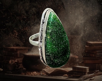 May include: A silver ring featuring a large, teardrop-shaped, deep green gemstone with a textured, sparkling surface. The metal band is simple and thin, while the setting has a decorative, slightly darkened edge surrounding the stone.