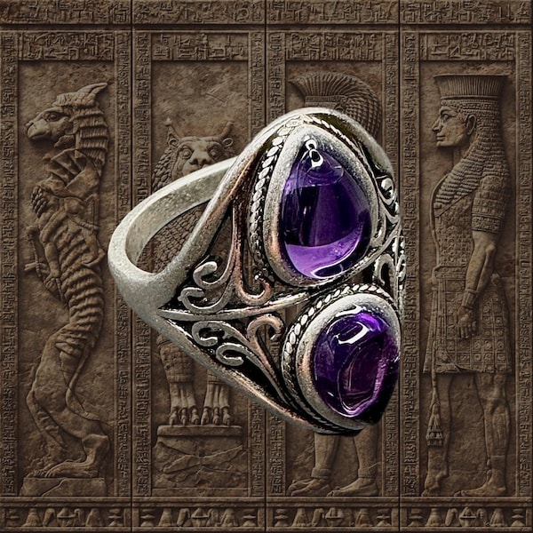 Puede incluir: Anillo de plata con dos grandes piedras preciosas ovales de color púrpura intenso. El anillo tiene un diseño ornamentado de estilo antiguo con intrincados trabajos en metal. El fondo es una superficie marrón texturizada con figuras egipcias antiguas talladas.