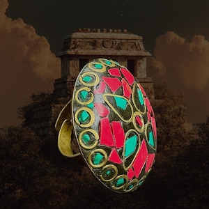 May include: An oval-shaped ring with a mosaic design. The ring features red, black, and turquoise-colored stones set in a gold-colored metal. The ring's design is reminiscent of ancient art. The ring is a statement piece.