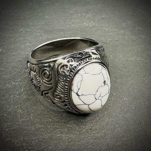 May include: A silver-toned ring with an oval, white stone featuring black veining. The ring's band is intricately detailed with embossed patterns. The ring is a bold statement piece.