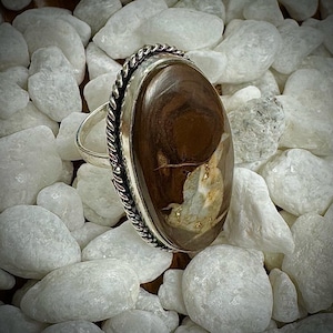 May include: A silver ring with an oval, brown gemstone. The gemstone has a unique pattern with lighter brown and white inclusions. The ring's setting features a twisted rope design around the stone, and it sits on a bed of white stones.