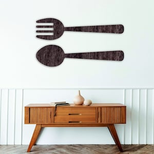 以下が含まれることがあります： 濃い茶色のフォークとスプーンのシルエットが描かれた木製壁飾り。壁飾りは、白い壁に、2つの引き出しが付いたミッドセンチュリーモダンスタイルの木製ドレッサーの上に設置されています。