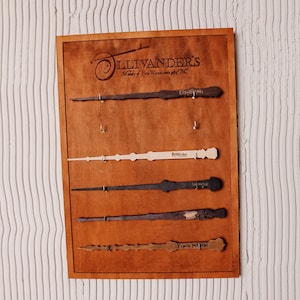 May include: A rectangular wooden display board featuring the Ollivander's Makers of Fine Wands since 382 B.C. logo at the top. Five distinct wands are mounted horizontally on metal hooks, each engraved with a unique spell name in dark lettering.