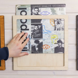 May include: A wooden frame with a white background holds a vinyl record. The record cover features a colourful design with a band name and album title in Russian. The record is being held by a hand.
