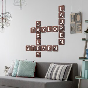 May include: A wall-mounted crossword puzzle display featuring dark brown wooden tiles with white letters. The tiles are arranged to spell the names Taylor, Carly, Steven, and Lauren, with each tile displaying a small numerical value in the corner.