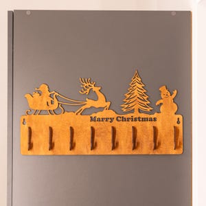Puede incluir: Perchero de madera con temática navideña con un Papá Noel en un trineo tirado por un reno, un árbol de Navidad y un muñeco de nieve. El perchero tiene ocho ganchos y el texto "Marry Christmas" está escrito debajo de las figuras.