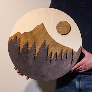 May include: A circular wooden wall art piece featuring a layered landscape design. The artwork displays a light wood sky with a circular sun, a medium-brown mountain peak, and a dark-stained foreground layer of detailed pine tree silhouettes.
