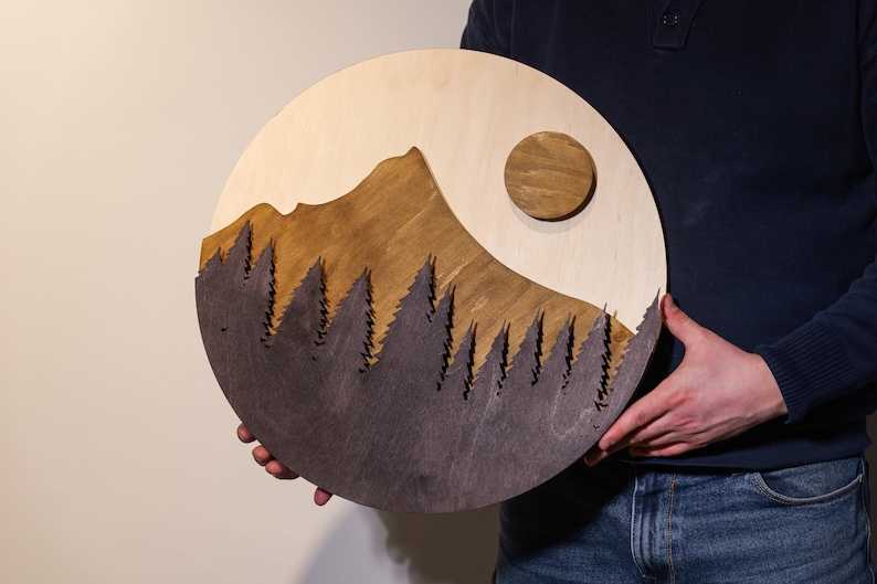 May include: A circular wooden wall art piece featuring a layered landscape design. The artwork displays a light wood sky with a circular sun, a medium-brown mountain peak, and a dark-stained foreground layer of detailed pine tree silhouettes.