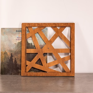 May include: A wooden geometric wall decor with a criss-cross pattern. The decor is brown and has a natural wood finish. It is displayed on a shelf with a book behind it.