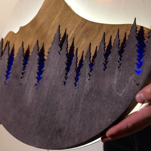 May include: A circular wooden wall art piece featuring a layered mountain and forest silhouette. The dark brown, laser-cut pine tree design is set against a lighter wood background, with vibrant blue LED lighting illuminating the gaps between trees.