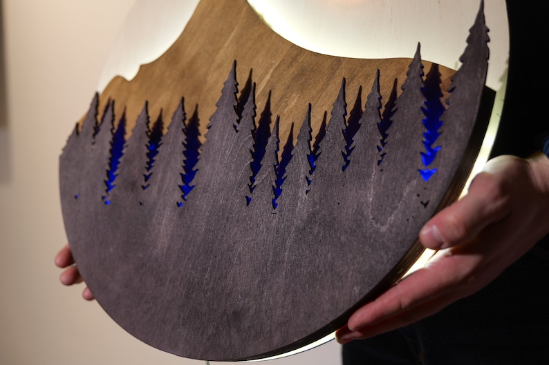 May include: A circular wooden wall art piece featuring a layered mountain and forest silhouette. The dark brown, laser-cut pine tree design is set against a lighter wood background, with vibrant blue LED lighting illuminating the gaps between trees.