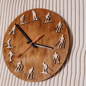 Può includere: Un orologio in legno con sfondo marrone e lancette nere. Il quadrante dell'orologio presenta un design di sciatori in silhouette attorno al quadrante.