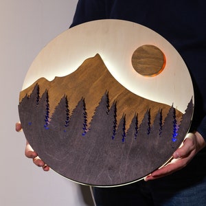 May include: A circular wooden wall art piece featuring a layered mountain landscape with a dark brown forest silhouette in the foreground and a light wood mountain peak behind it. A circular moon cutout glows with warm light, and blue light illuminates the trees.